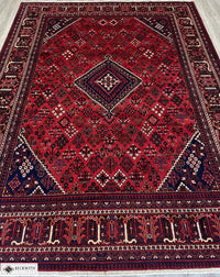 A rectangular, handcrafted wool rug with a geometric pattern, predominantly in red and blue colors, featuring intricate designs and a cotton foundation.