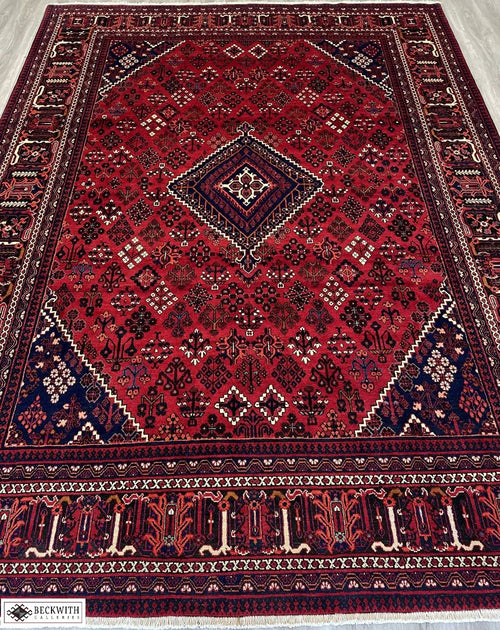 A rectangular, handcrafted wool rug with a geometric pattern, predominantly in red and blue colors, featuring intricate designs and a cotton foundation.
