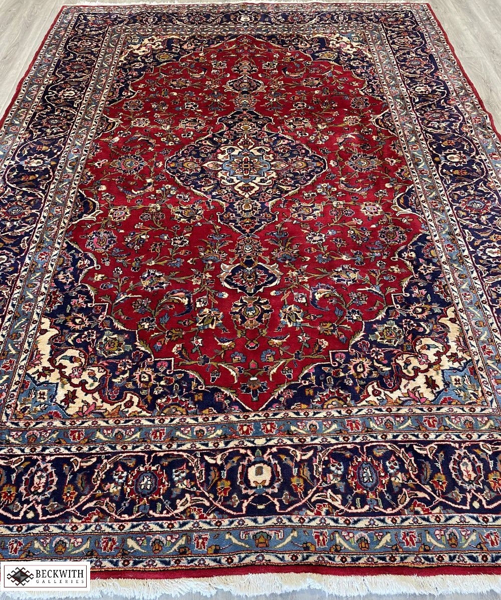 A Kashan-style rug with a floral pattern in blue, red, and gray hues on a white background.